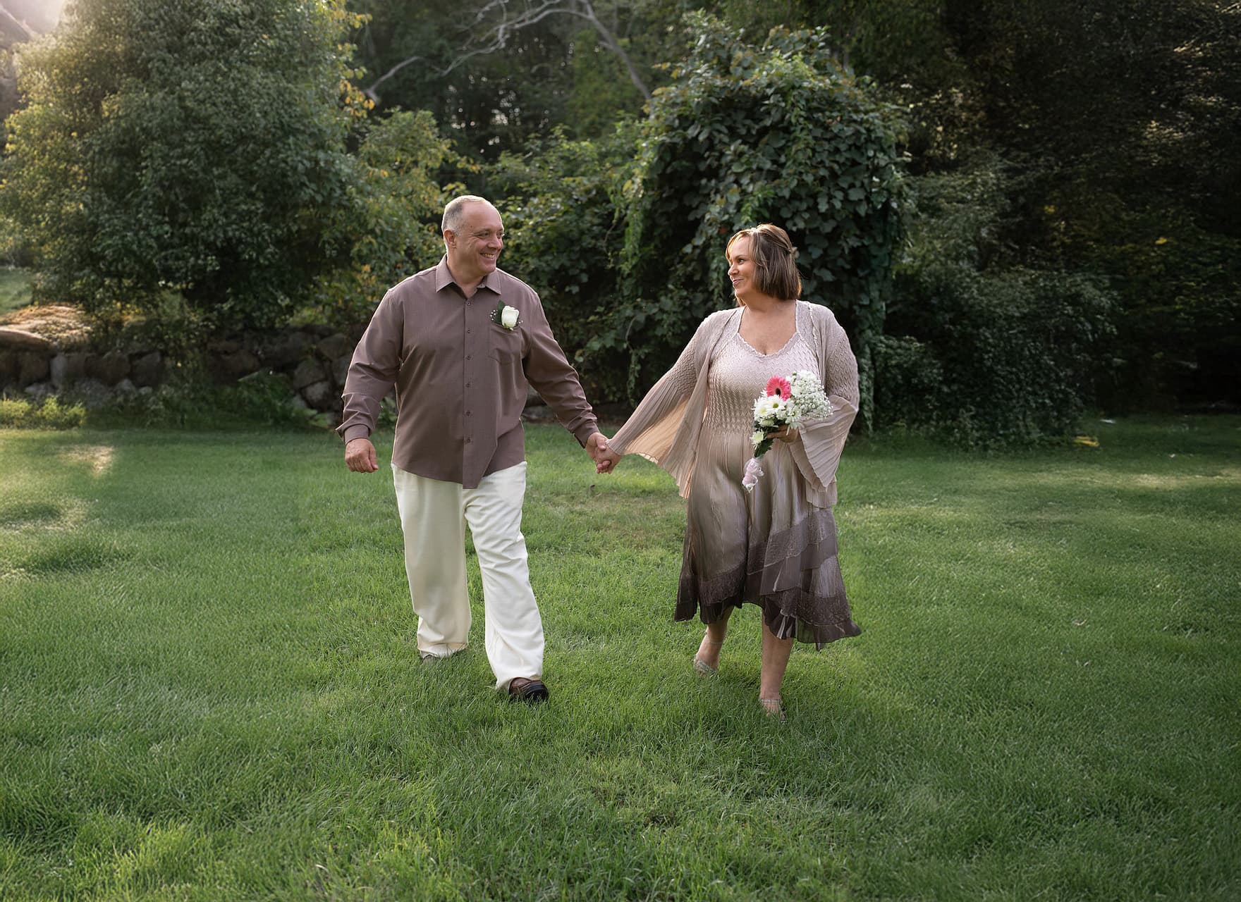 A couple walks hand in hand through a grassy area, smiling at each other.