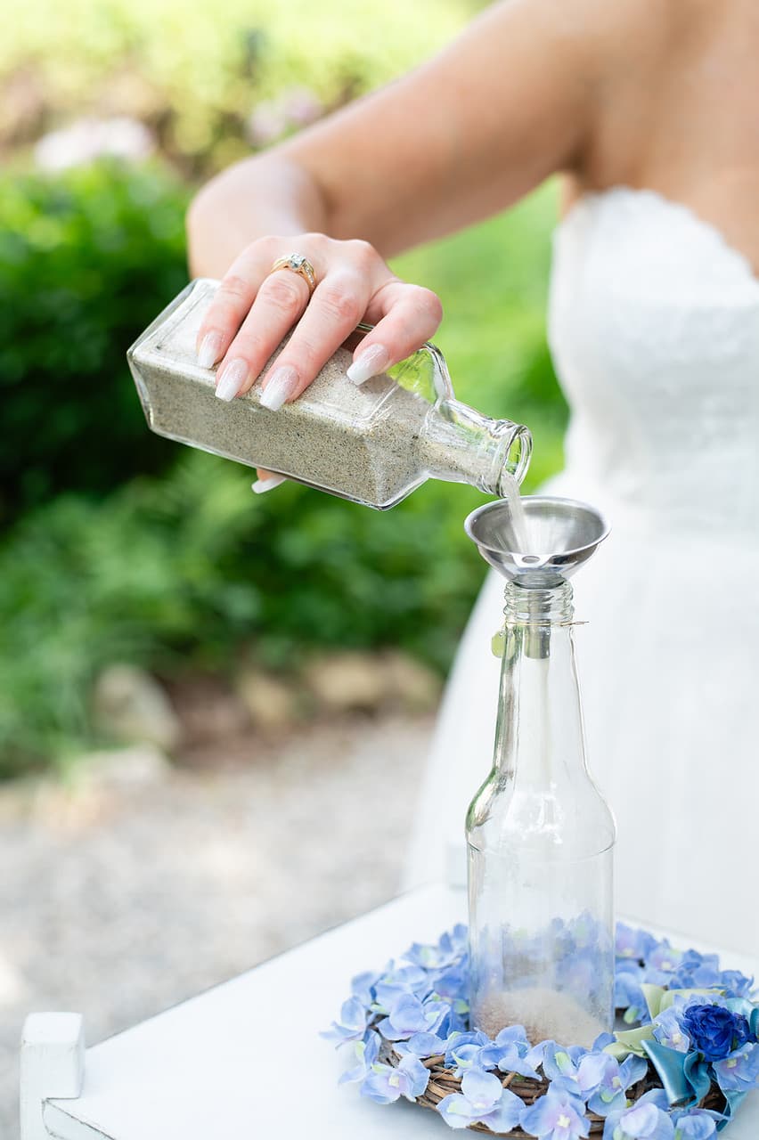 A person in a wedding dress pours sand from a glass bottle into a funnel above another glass bottle, surrounded by blue flowers.
