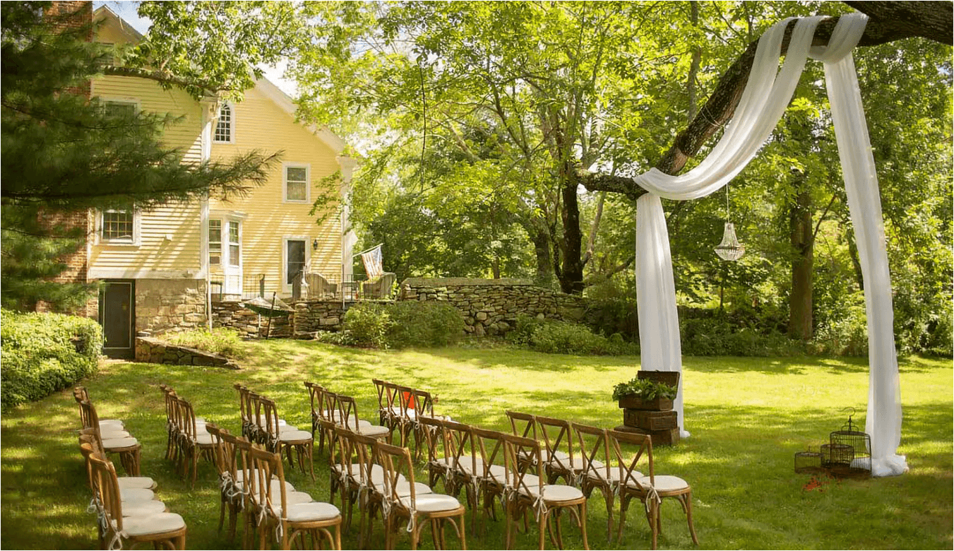 A charming outdoor wedding setup featuring rows of wooden chairs and a decorated archway beneath lush green trees by a historic yellow house. A charming outdoor wedding setup featuring rows of wooden chairs and a decorated archway beneath lush green trees by a historic yellow house.