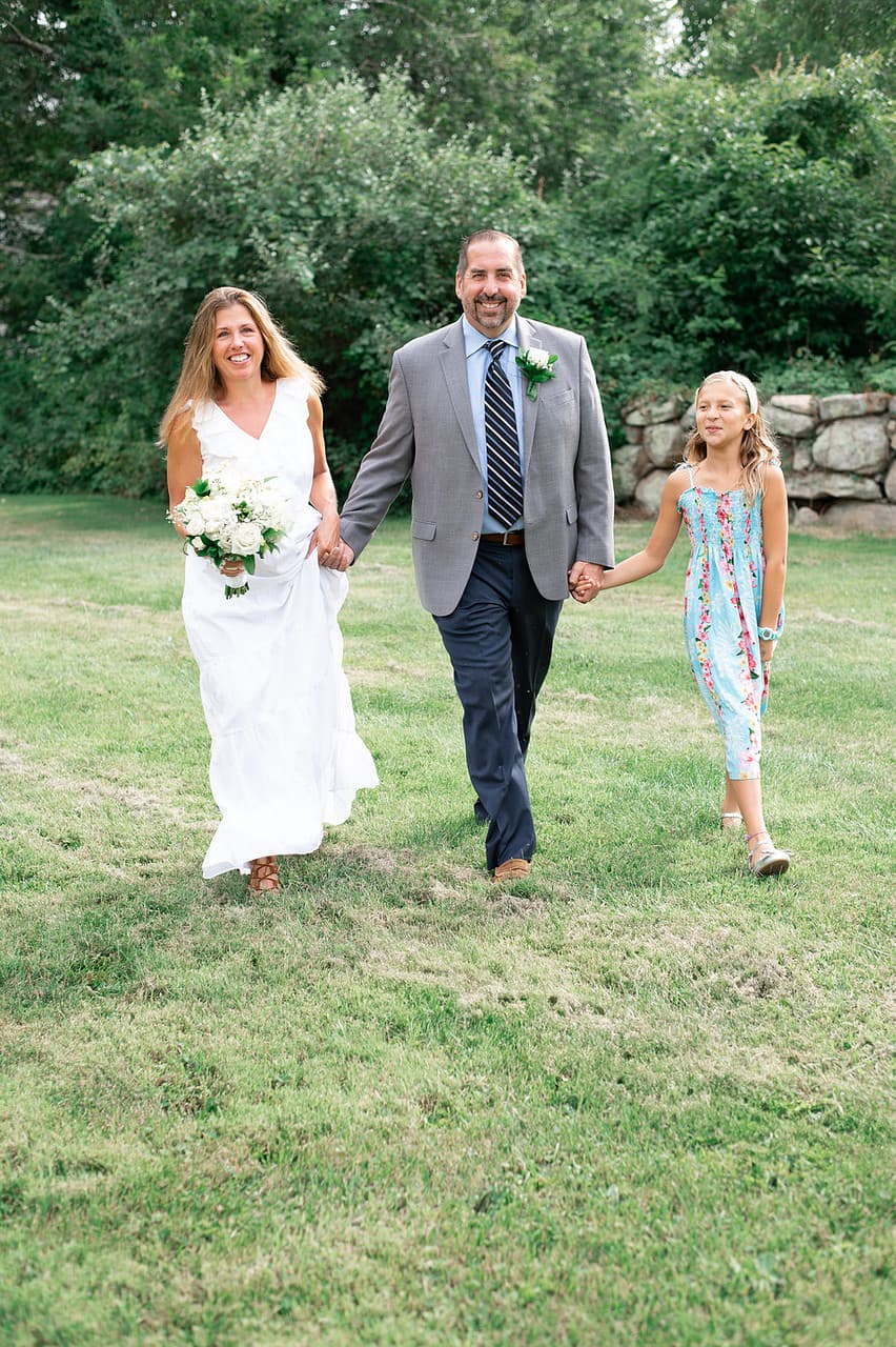 A bride, groom, and young girl walk hand in hand across a grassy field.