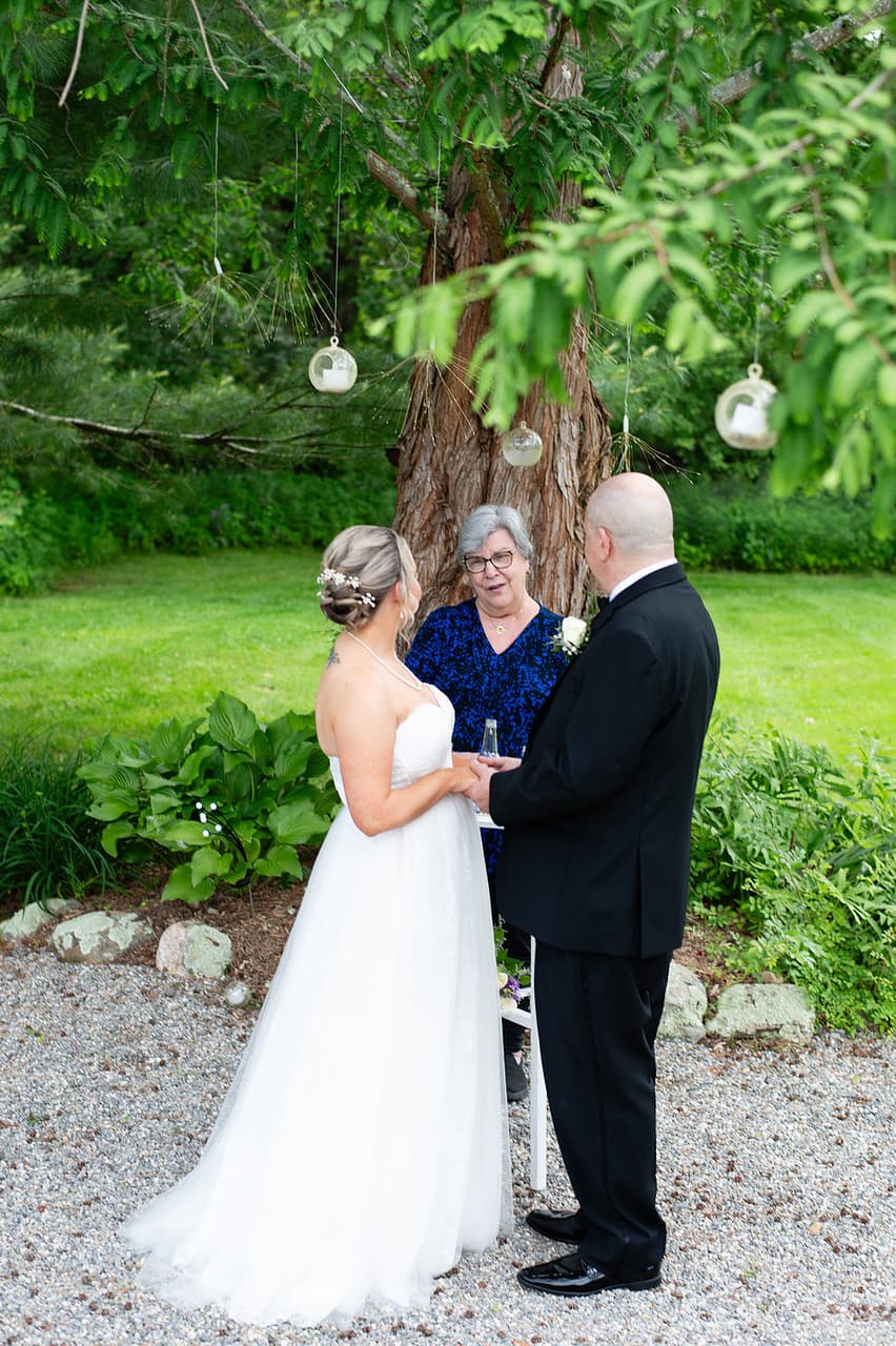 A bride and groom stand together under a tree while speaking with a woman in a blue dress, surrounded by greenery.