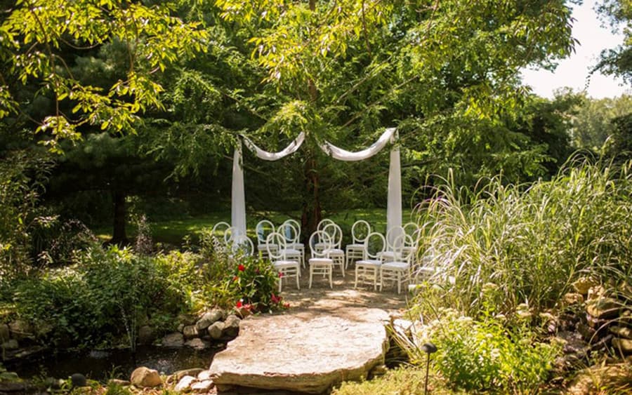 An outdoor wedding setup featuring white chairs and a draped canopy beneath a large tree, surrounded by lush greenery.