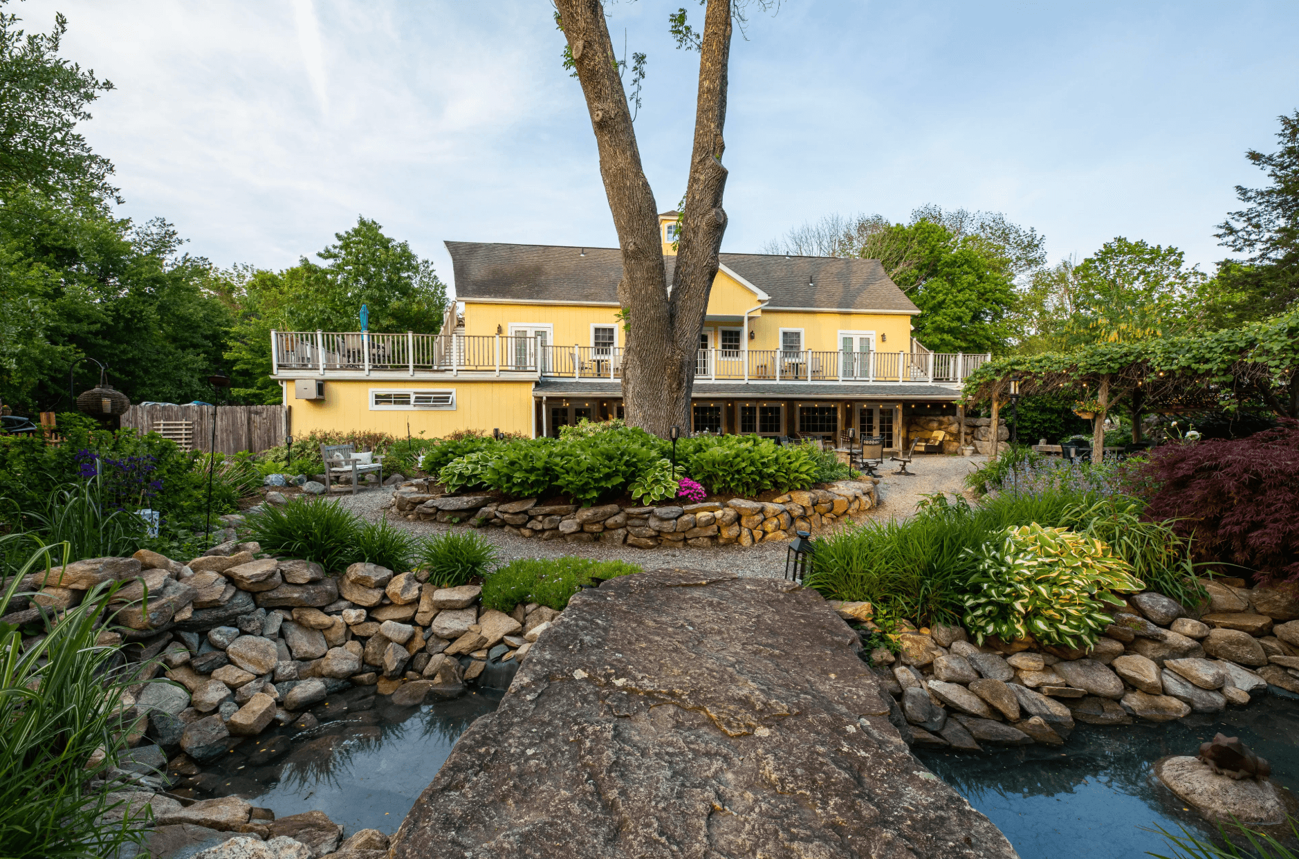 A yellow house with a deck surrounded by a landscaped garden and pond. A yellow house with a deck surrounded by a landscaped garden and pond.