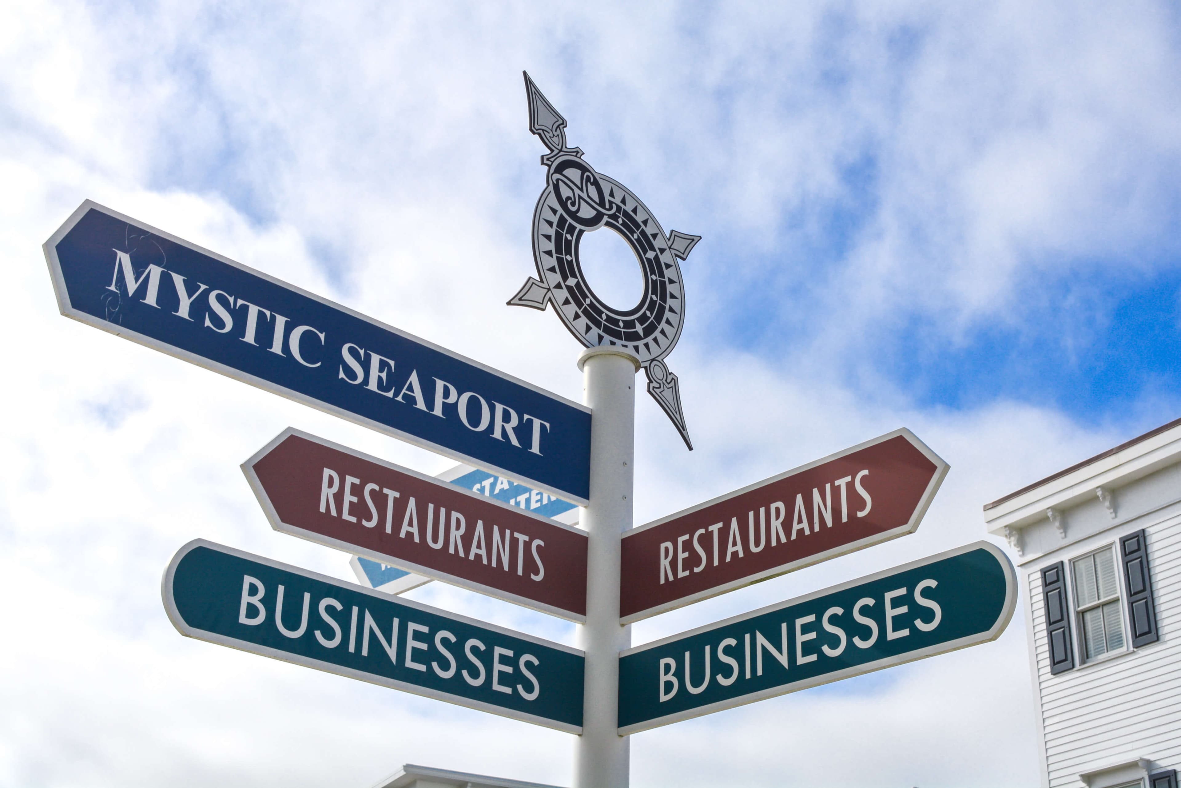 A directional signpost points to Mystic Seaport, restaurants, and businesses against a cloudy sky. A directional signpost points to Mystic Seaport, restaurants, and businesses against a cloudy sky.