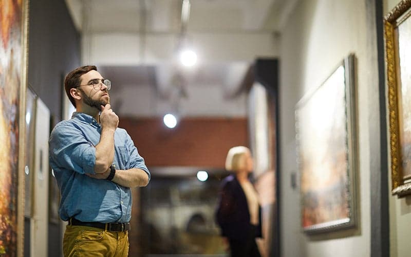 A man thoughtfully examines artwork in an art gallery while a woman observes nearby.