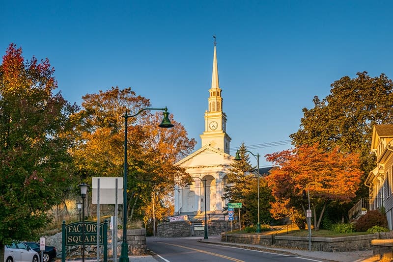 A white church with a tall steeple surrounded by autumn foliage on a clear day.