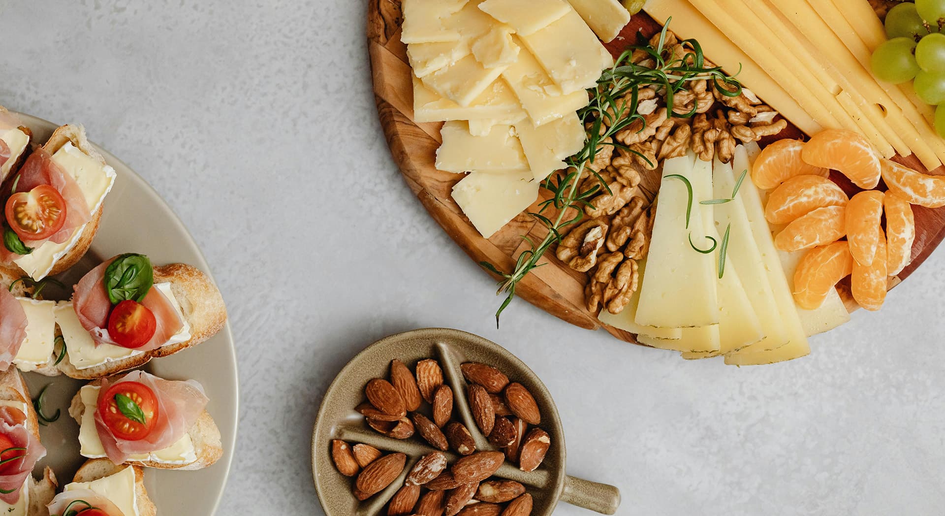 A spread of cheeses, nuts, and small sandwiches garnished with tomatoes on a light gray surface.