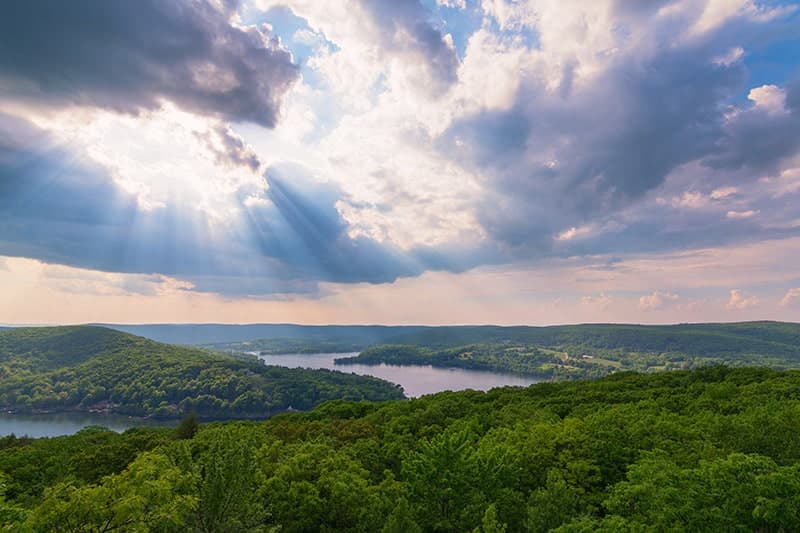 A panoramic view of lush green hills and a sparkling lake under a dramatic sky with rays of sunlight breaking through the clouds.