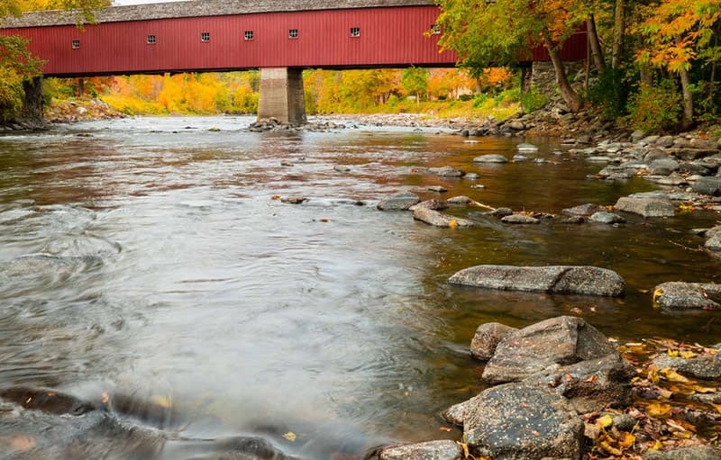 Red covered bridge over a flowing river surrounded by autumn foliage.